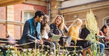 Studierende sitzen auf einer Bank am Campus Neues Palais und unterhalten sich. Eine Studentin hat einen Laptop auf dem Schoß.